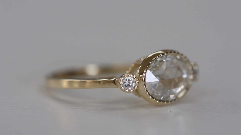 14k yellow gold custom engagement ring with oval cut white diamond center stone and two white side diamonds with milgrain detail