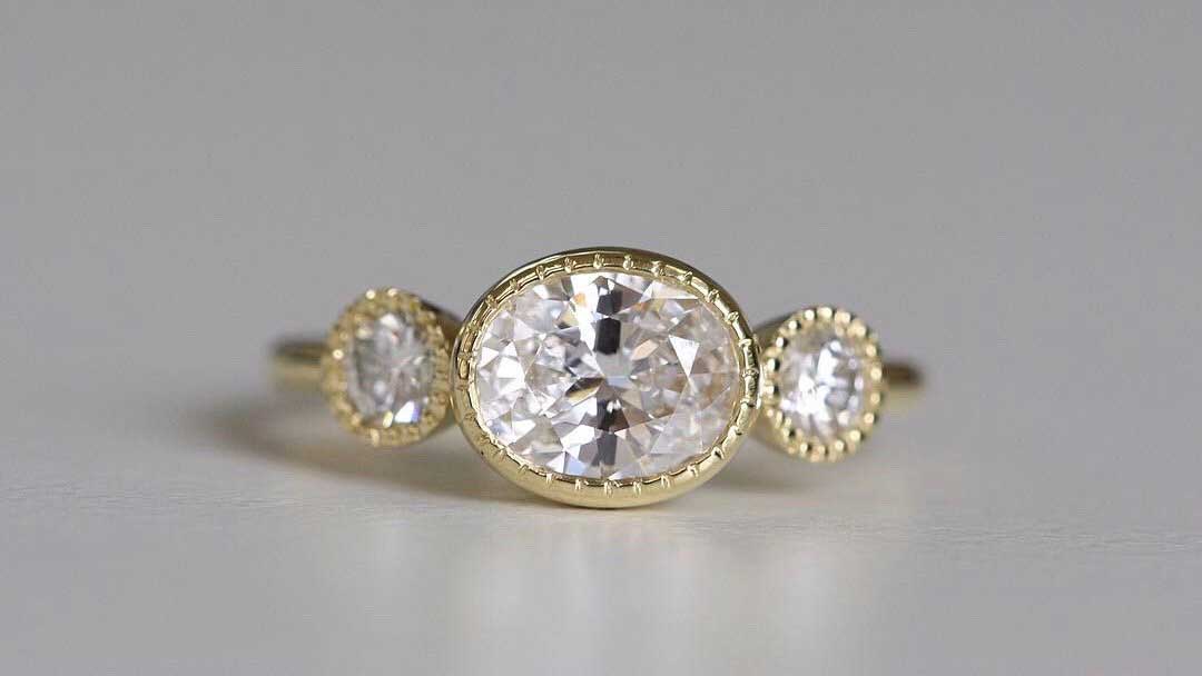 14k yellow gold custom ring with oval cut white diamond center stone and two white side diamonds with milgrain detail