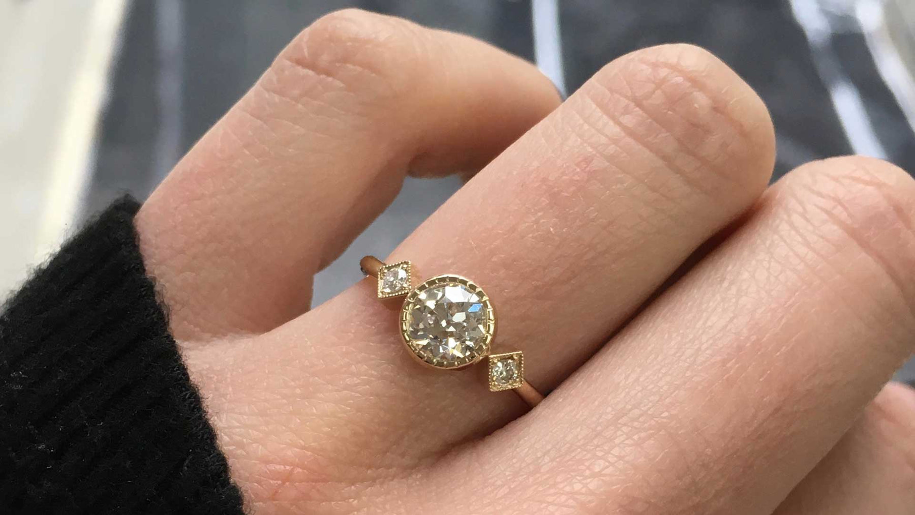 14k yellow gold custom engagement ring with white diamond center stone and two side white diamonds and milgrain detail on hand