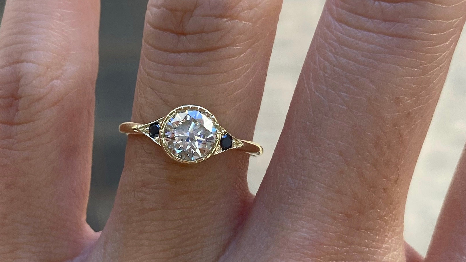 14k yellow gold custom engagement ring with round cut white diamond center stone with blue sapphire side stones and milgrain detail on hand